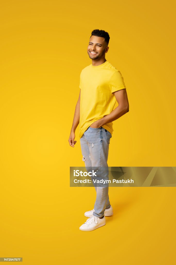 Pleased indian man in casual wear stands isolated on yellow, male student, young handsome guy standing Pleased indian man in casual wear stands isolated on yellow, male student, young handsome guy standing in relaxed pose and looking aside, holding hand in pocket of jeans, full length Men Stock Photo