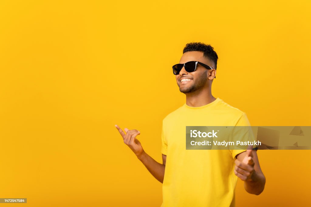 Excited positive young arab man in sunglasses rejoicing, raising hands up and having fun, good mood. Indoor studio shot isolated on orange background Excited positive young arab man in sunglasses rejoicing, raising hands up and having fun, having good summer mood. Indoor studio shot isolated on yellow background Sunglasses Stock Photo