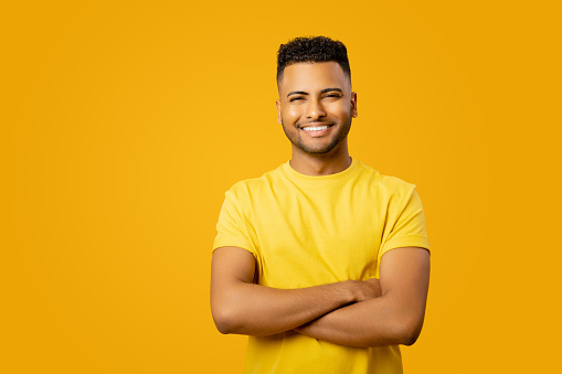 Smiling confident Indian guy wearing casual t-shirt stands with arms crossed isolated on yellow, studio shot of cheerful latin businessman, success concept, copy space