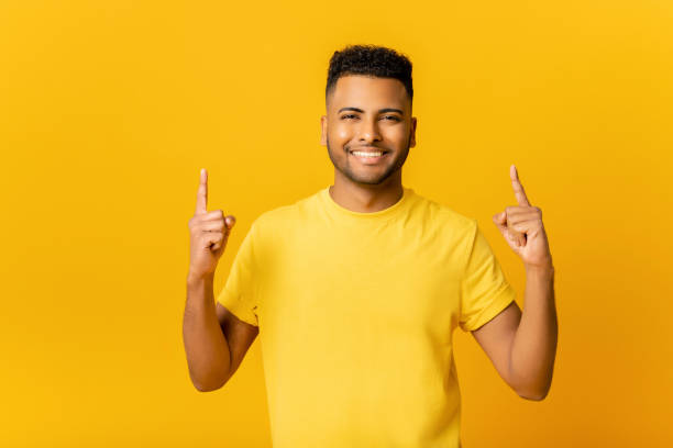 istock Cheerful overjoyed Indian guy points fingers up at empty copy space, isolated on yellow background 1472547657