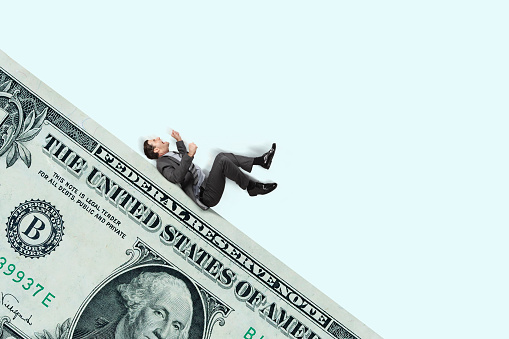 A man on his back slides down a financial slippery slope in front of a light blue background.