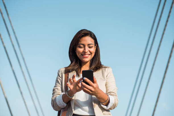 A happy elegant young latin woman stands and uses her phone to type an important message. A businesswoman using technologies outdoors. A happy elegant young woman stands and uses her phone to type an important message. A businesswoman using technologies outdoors. mediterranean woman portrait stock pictures, royalty-free photos & images