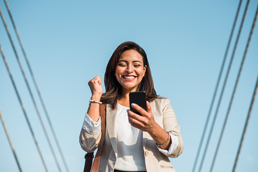 portrait of a businesswoman smiling happily with a fist in the air because she has won an award by looking at her mobile phone.