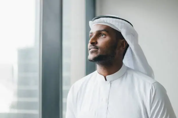 Portrait of Successful Muslim Businessman in Traditional White Outfit Standing in His Modern Office, Using Smartphone Next to Window with Skyscrapers. Young Saudi, Emirati, Arab Businessman Concept.
