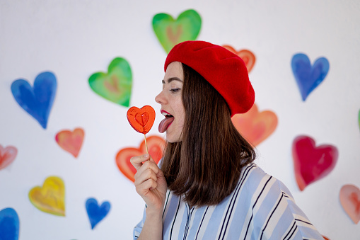 a girl against a background of colorful bright hearts licks a big lollipop in the form of a heart with her tongue in red, the concept of valentine's day, love