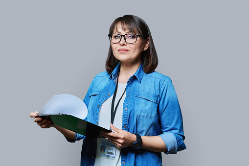 Portrait of woman worker with name identity card of industrial center clipboard, on grey color background. Confident female engineer manager supervisor auditor inspector expert looking at camera