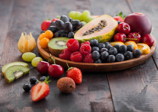 Fresh raw organic summer berries and exotic fruits in round wooden plate on dark wooden kitchen background. Papaya, grapes, nectarine, orange, raspberry, kiwi, strawberry, lychees, cherry. stock photo