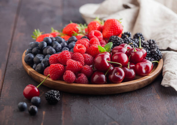 las bayas frescas orgánicas de verano se mezclan en una bandeja redonda de madera sobre el fondo de la mesa de madera oscura. frambuesas, fresas, arándanos, moras y cerezas con paño de cocina de lino. espacio para texto - baya fotografías e imágenes de stock