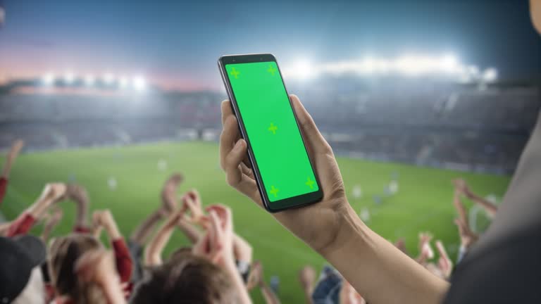 Sport Stadium Championship: Person's Hand Holding Green-Screen Chroma Key Smartphone. Sports Match with Fans on Tribune Cheering for Favourite Team to Win. Isolated POV Close-up Copyspace Template.