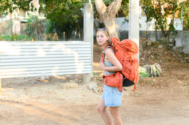 Side view of young lady woman blonde with long hair orange backpack in casual summer outfit looking at camera while walking during trip in Central America