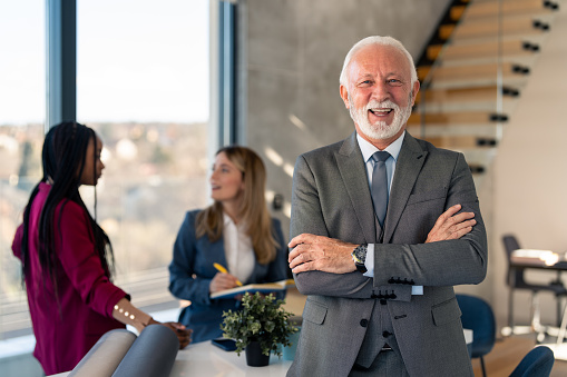 Portrait of smiling confident senior businessman leader wearing suit looking at camera with arms crossed standing in office at diverse team meeting.