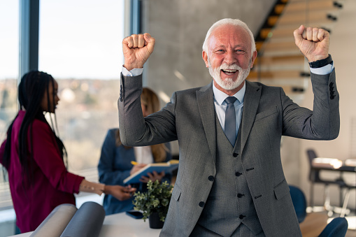 Portrait of successful senior corporate businessman in suit at modern office cheering after successful professional achievement financial deal with business partners at work.