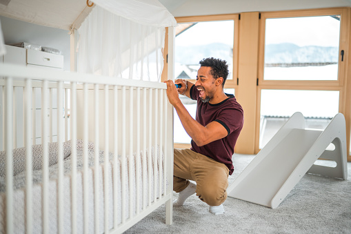 Cheerful mid adult Latin American man building furniture in a baby room. He is happy and excited for the arrival of his first daughter.