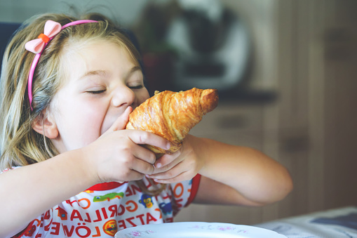 Smiling child at breakfast. Food and happy kids. The girl is eating a croissant. Cute preschool girl having healthy meal