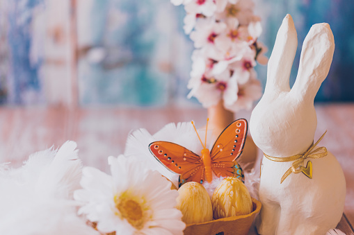 Close up of gold painted Easter eggs with a branch of almond blossoms, a white daisy, an orange butterfly and a white Easter bunny on a wooden table on blue vintage background. Creative color editing with added grain. Very soft and selective focus. Part of a series.