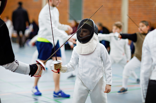 Little kid boy fencing on a fence competition. Child in white fencer uniform with mask and sabre. Active kid training with teacher and children. Healthy sports and leisure