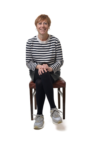 front view full length portrait of a woman in skirt, striped sweater and sneakers sitting on chair and legs crossed and smilling on white background
