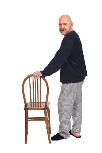 side view of a man in pajamas standing plaiyng with chair and looking at camera on white background