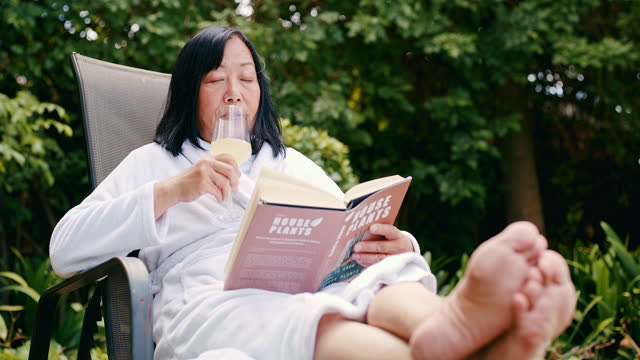 Relax, book and wine with old woman in garden for peace, holiday and retirement at resort. Summer, nature and lounge with asian lady and champagne at hotel for free time, reading and vacation