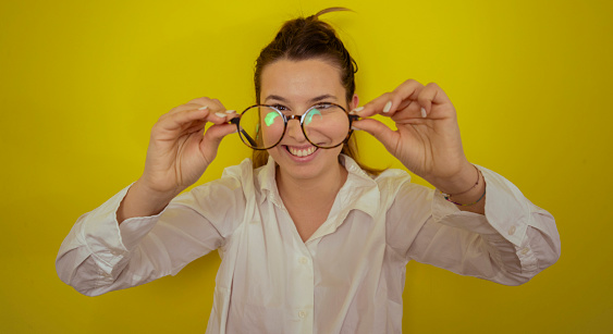 Smiling woman holding eyeglasses while standing in front of yellow background