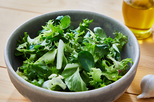 Delicious homemade gourmet healthy green salad bowl, with olive oil, garlic and goat cheese in olive oil, served in a rustic handmade ceramic bowl, with seasoning and herbs on a natural wooden kitchen or restaurant table, representing a healthy lifestyle, wellbeing and body care, top view with a copy space