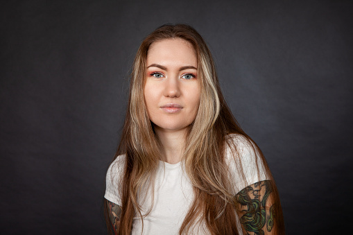 Studio portrait of a young white woman with long brown hair, with a tattoo, in white t-shirt, against a black background