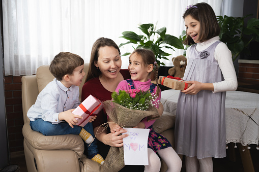 Mother is having a happy mother's day with her children surprising her and giving bouquet of flowers and gifts.