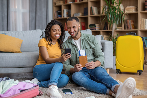 Loving african ameican couple booking tour or hotel online, using mobile phone, holding passport with tickets, contacting tourist agency on web, sitting on floor in living room