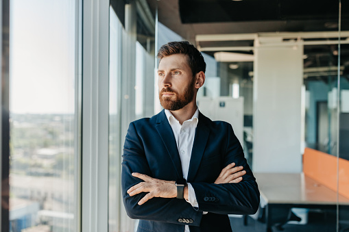 Successful businessman in formal wear standing with folded arms in modern coworking office and looking through window, thinking about startup, planning new projects, waiting for meeting