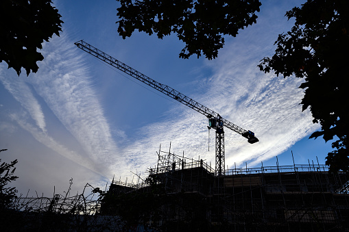 Silhouette of a construction crane on a building site. Crane is framed by trees and set against a background of a blue sky with white clouds.
