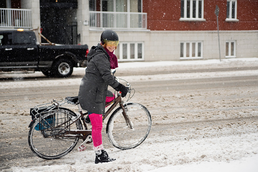 Athletic 50+ woman commuting with bicycle in of the winter snow. She has red hair and is dressed in bright pink and dark gray. Horizontal full lenght outdoors shot with copy space. This was taken in Montreal, Quebec, Canada.