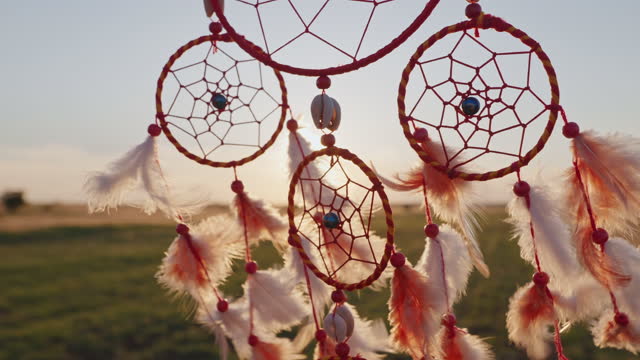Dream catcher with orange feathers close up rotate in mountains, swaying with light wind slow motion agains blue sky at sunset in summer. Amulet boho style, Indian style. Bright disk of sun, rays sun