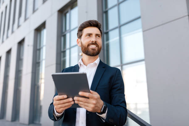 istock Happy confident handsome young caucasian man with beard in suit typing on tablet, look at empty space 1472661323