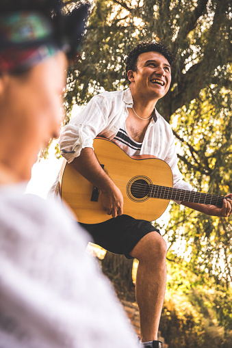 Low angle view of smiling young man having fun and enjoying playing the guitar and singing for his girlfriend while they are out on a picnic.