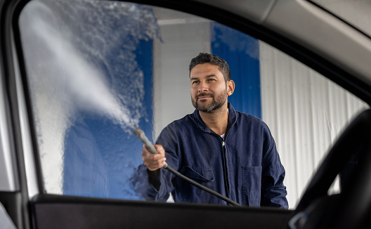 Latin American man working at the car wash cleaning a window with a high-pressure hose - small business concepts