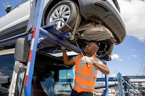 African American man towing a vehicle on a car transporter and adjusting the straps - freight transportation concepts
