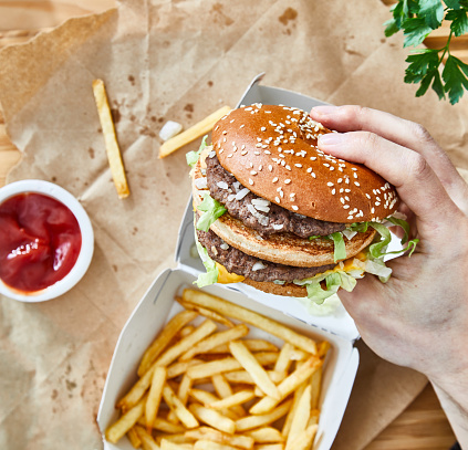 Man holding a fresh, tasty and delicious hamburger with French fries on a recycling paper, with ketchup and cola, served on a bamboo wooden cutting board, bar, restaurant or home kitchen table, top view with copy space, representing fast food and city life