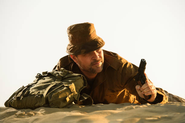 A soldier in uniform holding a pistol lying on a dune. Low angle view, landscape format, shot for book cover purposes. Warm-toned, blank space above extendable for copy space.