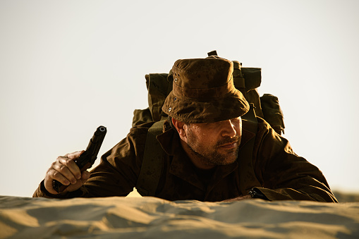 A rugged-looking soldier in uniform holding a pistol lying in sand on a dune. Low angle view, landscape format. White space behind extendable for portrait format book cover purposes, toned.