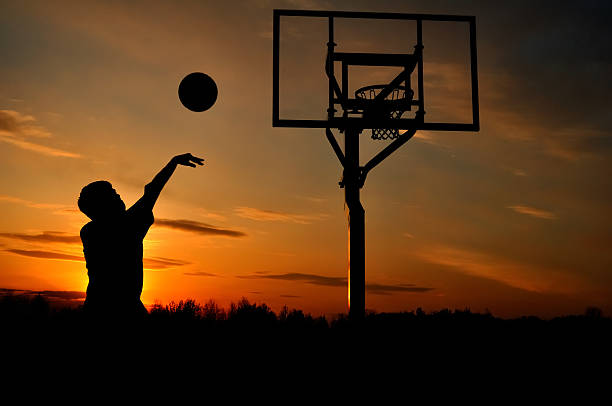 Silhouette of a teenage boy shooting a basketball at sunset stock photo