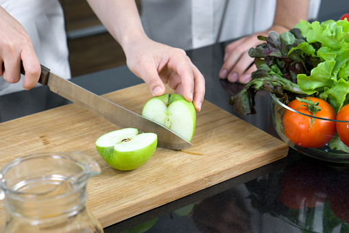 Closeup hand holding knife cutting green apple on a wooden chop board. A glass bowl with a variety of vegetables is placed on the kitchen counter.