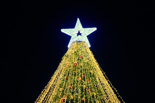 Christmas tree with big white star topper decorated yellow garlands and colorful decorative bulbs on night blue sky background, outdoor holiday atmosphere. Festive Christmas tree with yellow lights