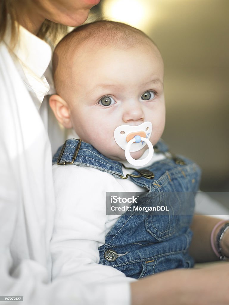 Baby With Pacifier in Mother's Lap Portrait of a baby girl with a pacifier, sitting in her mother's lap and looking at the camera. Vertical shot. Adult Stock Photo