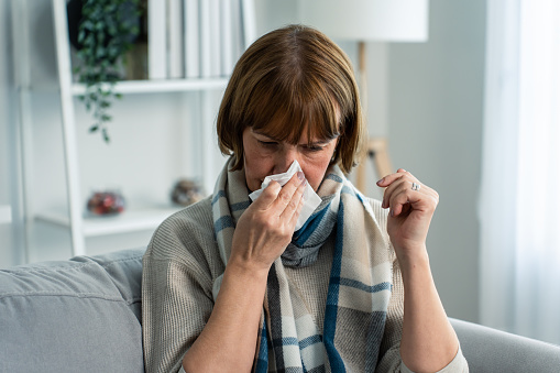 Caucasian senior woman having running nose and sneezing in living room. Attractive elderly female sitting on sofa feel bad and suffer from allergy then put tissue cover her nose while sneez in house.