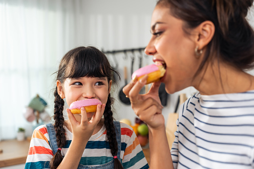 Caucasian happy family, parent spend time with kid together at home. Adorable young girl daughter eatting sweet donut with mother with happiness in kitchen. Activity relationship in house concept.
