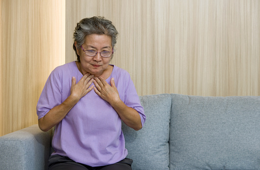 Asian elderly with eyeglasses, 
place both hands on the chest due to nausea while sitting on the sofa in the living room.