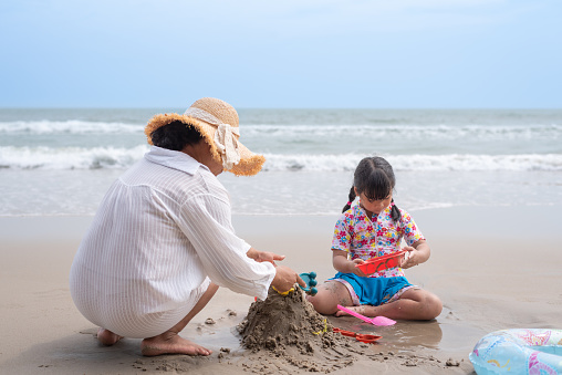 Wide shot of Asian mother playing with pre-school daughter at beach, making sand castle. Child care, family, mother and daughter, babysitting concept.