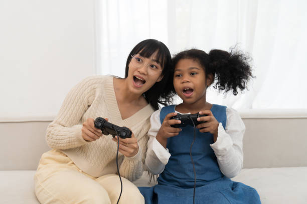 週末に一緒にリラックスするために家でビデオゲームをする若い母と娘。家のソファで子供とジョイスティックゲームをする幸せなアジアのお母さん - group of people people family multi ethnic group ストックフォトと画像