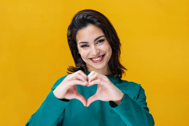 istock Romantic happy woman making heart shape with hands while smiling to camera isolated on yellow 1472745751