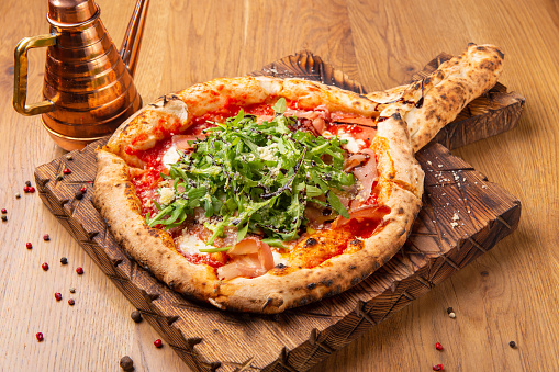 Oven baked pizza with mozzarella cheese, dried ham, tomatoes, balsamic vinegar and arugula over wooden background. Close up view